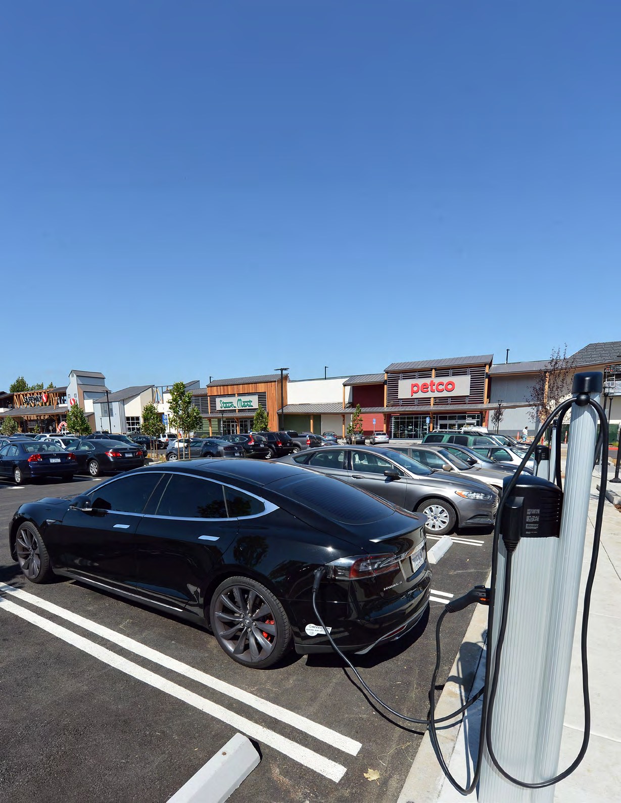 Black Tesla Model S plugged into a charging station at a shopping center parking lot with Petco and other retail stores visible in the background